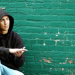 Color photo of a young, hispanic man in an alley crouching by a green brick wall thinking about his life on the street as a gang member.